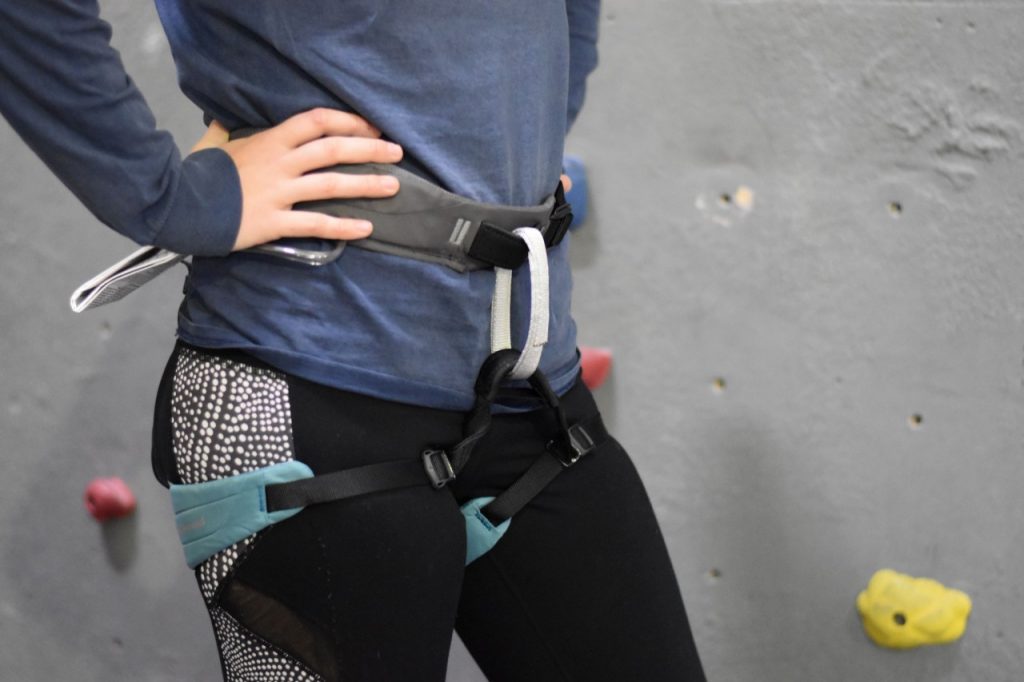 A woman models Black Diamond women's Momentum harness. The leg straps are teal and adjustable, the waist strap is grey, and the belay loop is silvery white.