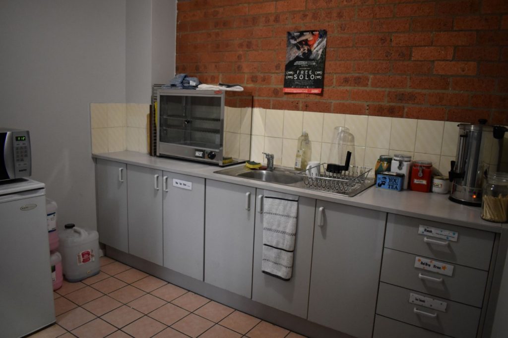 A photo of our gyms kitchen, showing a mini fridge with a microwave sitting on top of it, a pie warmer, a sink, and a large urn full of hot water with implements for tea and coffee. All of the cupboards are labelled with their contents, including mugs, plates, cutlery, etc.