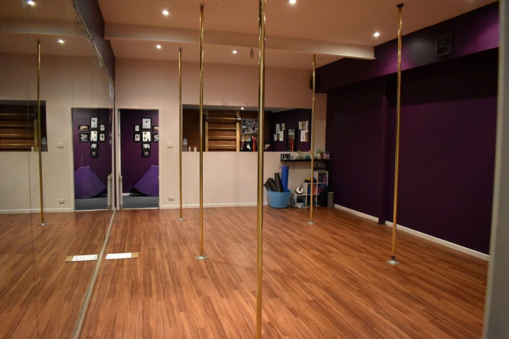 The walls of the pole studio are painted purple and white, and the floor is wood. Full-length mirrors cover two walls. There are six golden poles. A lounge area can be seen in the background.
