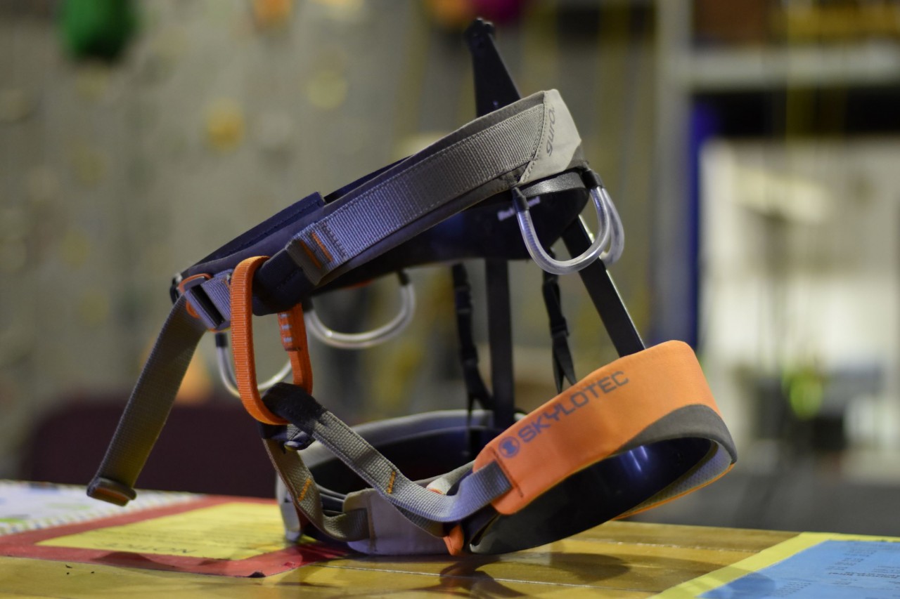 A women's Skylotec Guro harness positioned on a display frame. The harness is grey with orange leg straps, belay loop, and detailing.