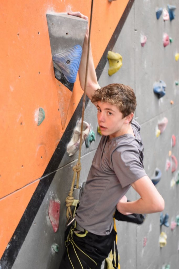 A young climber leans back on one arm, his hand gripping the top of a feature in the middle of a climb, and reaches into his chalk bag with his other hand
