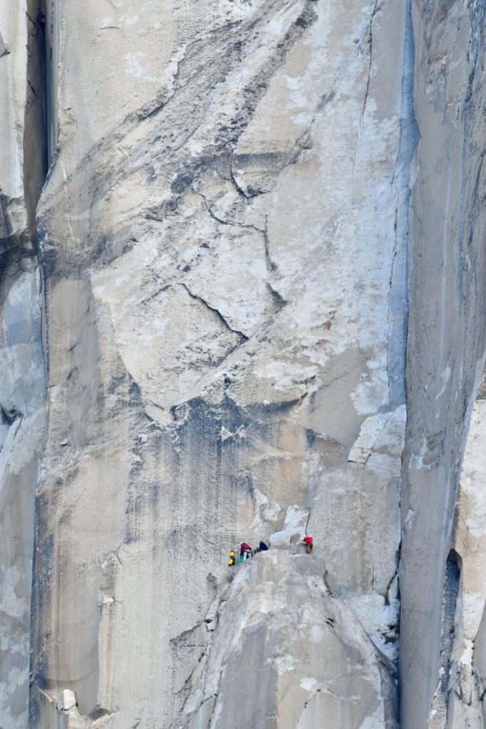 A photo taken of El Cap from the meadow below, showing a massive granite face with three small figures - Cait, Greg, and Peter - on the side.