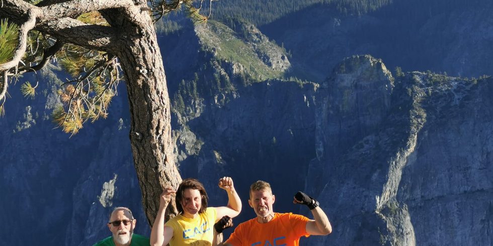 Peter, Cait, and Greg hold up fists and smile as they stand on the summit of El Cap together after topping out on the Nose Route. They stand beside a lone pine tree, and are all wearing Climbers Against Cancer t-shirts.