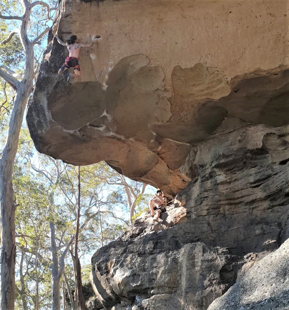 Kale lead climbs on an outdoor cliff, pulling himself over the overhang of a large roof. His belayer sits on a ledge beneath the roof, lead belaying and watching his progress.