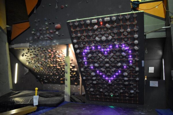 A photo of the Tension Board at Mitchell and the adjacent steep training cave. The Tension Board lights make a pink heart.