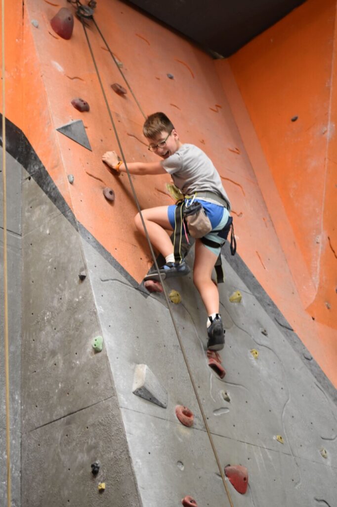 A photo of a young boy in our Geckos program climbing at Mitchell. He is glancing down at his belayer and smiling.